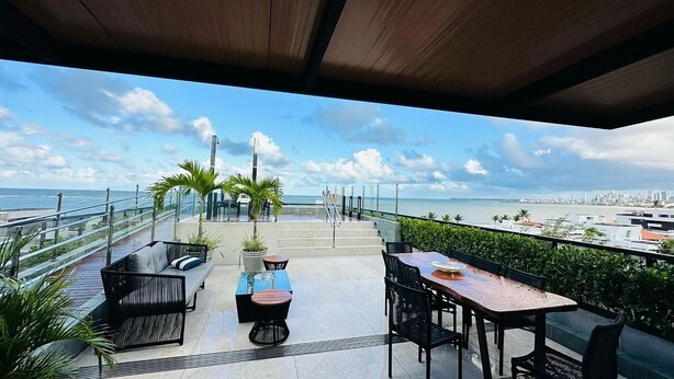 Een terras buiten met uitzicht op de oceaan, met een zithoek, tafels, stoelen, een trap en een balustrade.