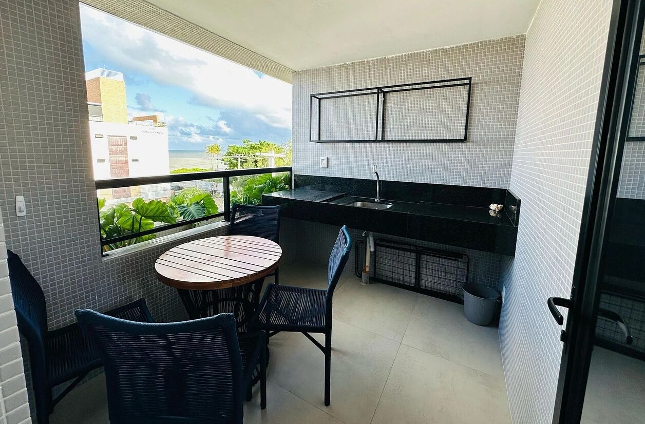 A compact balcony with a round table and chairs, a small kitchen area with a sink, and a view of buildings and greenery.