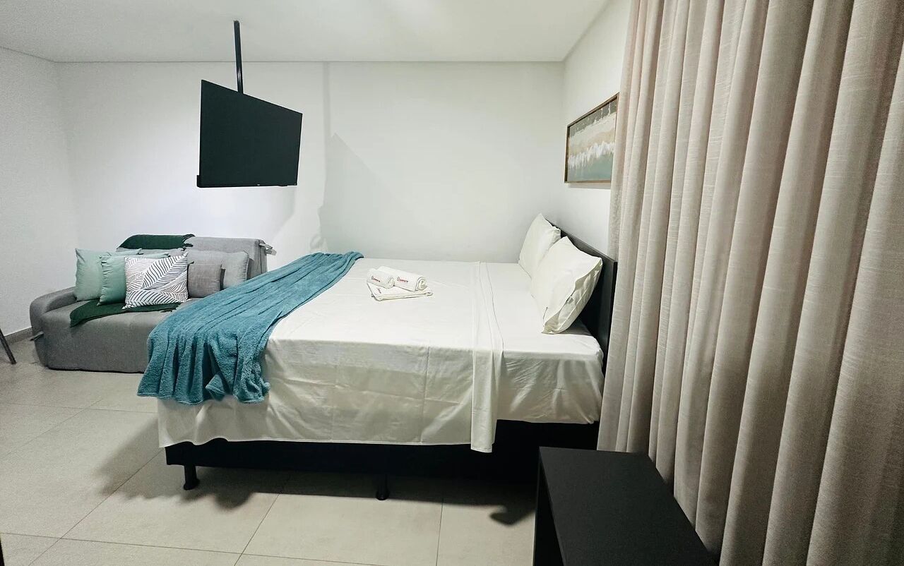 A bedroom with a bed, a bedside table, a chair, a TV, and a window with curtains.