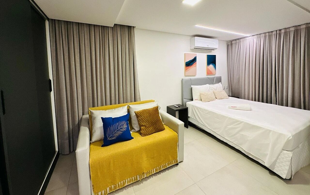 Una habitación de hotel con cama, sofá, mesita de noche y un ventanal con cortinas.