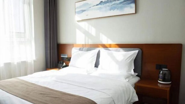 A hotel room with a bed, a nightstand with a coffee maker, a painting on the wall, and a window with curtains. - Home Inn Huayi (Linyi Lanling Yucai Road) (Lanling)
