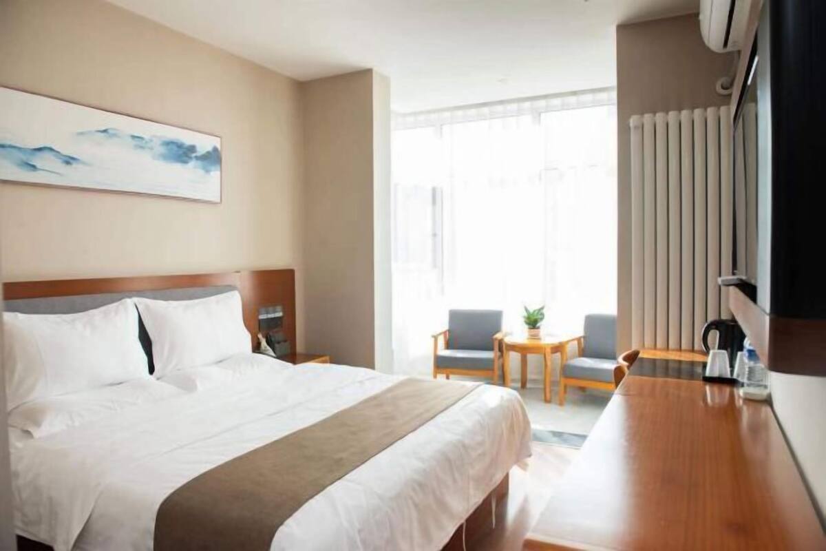 A hotel room with a bed, a desk, two chairs, a window, and a radiator.