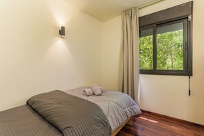 A single bed with a grey mattress and pillows, a wooden floor, a window with curtains, and a wall-mounted light fixture. - Warm Getaway 24/7 Power in Kfardebian (Jabal Lubnan)