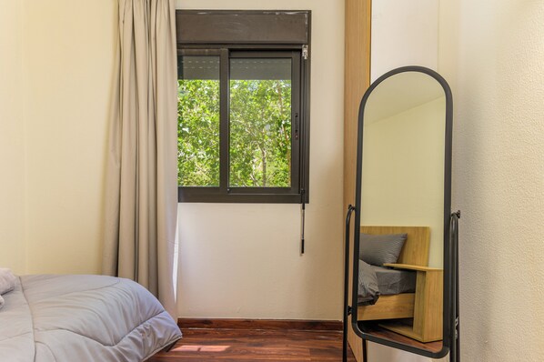 A bedroom with a bed, a window with curtains, a mirror, and a view of greenery outside.