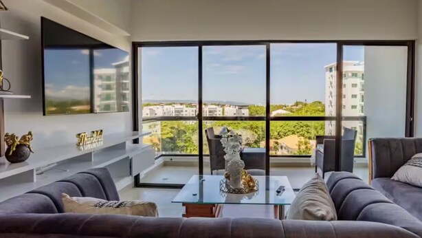 A modern living room with a large window, grey sofas, a glass coffee table, and a decorative vase.