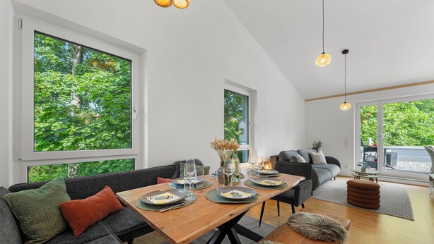 Una sala de estar moderna con una mesa de comedor de madera preparada para cuatro comensales, un sofá gris y un amplio ventanal con vistas a zonas verdes.