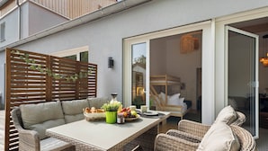 Un patio con mobiliario de mimbre, una mesa con frutas y una botella, y un ventanal corredizo que da a una habitación interior.