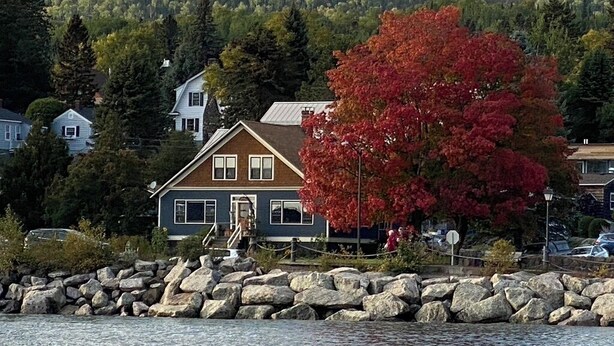 Une maison au bord d’un lac, avec une rive rocheuse, entourée d’arbres et d’une colline boisée en arrière-plan.