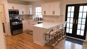 A modern kitchen with white cabinetry, stainless steel appliances, a central island with bar stools, and a black-framed door with windows. - Modern Mission Escape Home - Family Friendly - Close to All Attractions (Mission)