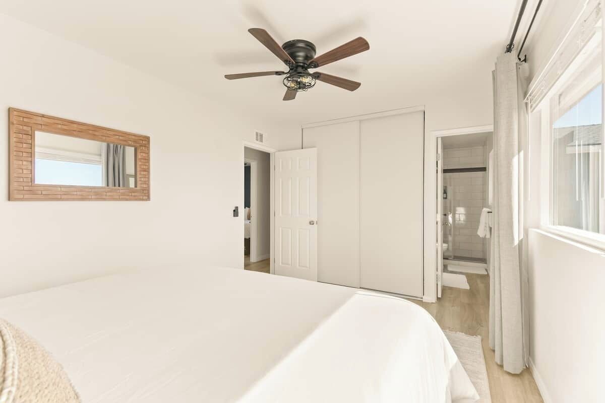 Una camera da letto con un letto, una ventola a soffitto, uno specchio, un armadio e un bagno parzialmente visibile attraverso una porta aperta.