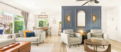 A modern living room with a dining area, a ceiling fan, and large windows offering a view of the outdoors.