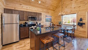 A wooden cabin interior with a kitchen featuring a stainless steel refrigerator, microwave, and stove, a wooden dining table with chairs, and a large window with a view of trees. - Hot Tub + Foosball Access to Pool, Tennis, Gym Chill'n in the Smokies (Gatlinburg)