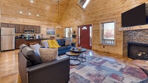 A spacious living area with a wooden ceiling, a fireplace, a sofa, a coffee table, and a kitchen in the background. - Hot Tub + Foosball Access to Pool, Tennis, Gym Chill'n in the Smokies (Gatlinburg)