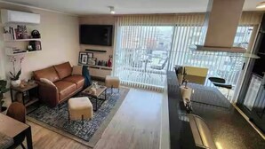A modern living room with a brown leather sofa, a coffee table, and a small side table with a plant. There is a large window with blinds, a flat-screen TV mounted on the wall, and a balcony with a chair.