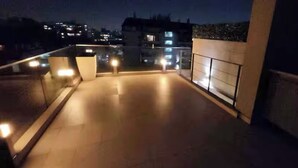 A rooftop terrace with a tiled floor, a railing, and a view of the cityscape at night.