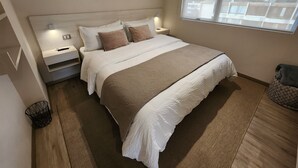 A modern bedroom with a large bed, two bedside tables, a window with a view of buildings, and a wooden floor.