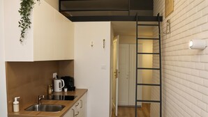 Ett kompakt kök med en bänkskiva i trä, en diskho i rostfritt stål och en kaffebryggare. En svart stege står lutad mot väggen, och en vit tegelvägg syns i bakgrunden.