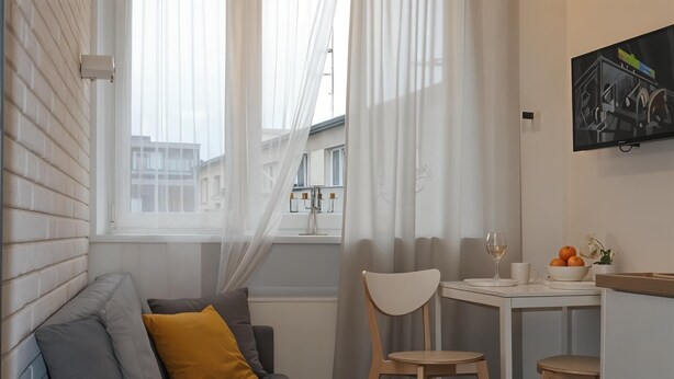 Ett modernt vardagsrum med en grå soffa, en gul kudde, ett vitt bord med två stolar, ett glas vin och en skål med apelsiner.
