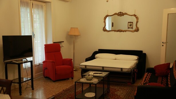 Una habitación de hotel compacta con una cama, un sillón rojo, una mesita, un televisor y una lámpara.