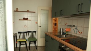 Una cocina compacta con encimera de madera, un lavamanos y una estufa. Hay dos sillas con cojines, un estante con objetos decorativos y una puerta con un panel de vidrio esmerilado.