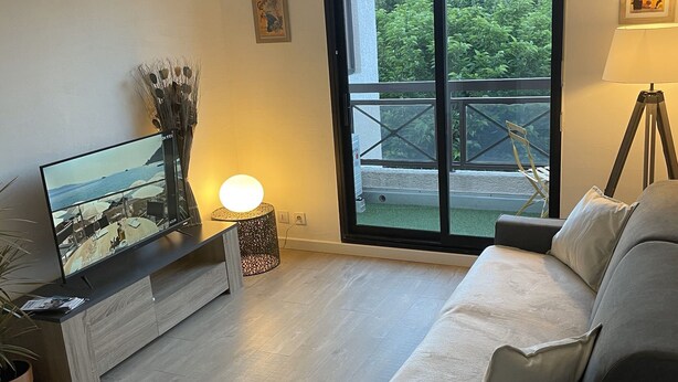 Een moderne woonkamer met een flatscreen televisie, een bank, een eettafel en een balkon met uitzicht op groen.