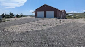 A single-story red building with two white garage doors, a gravel driveway, and a clear blue sky. - Charming 3-bedroom house with AC, WiFi in beautiful Lander (Lander)