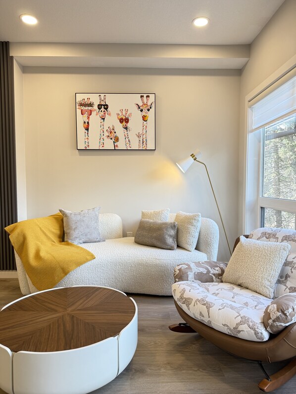 Uma sala de estar moderna com um sofá, poltrona e uma mesa de centro redonda. Há um quadro na parede e uma janela com persianas.