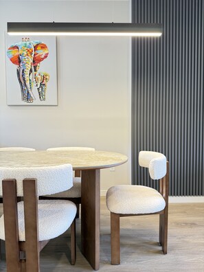 Uma área de refeições com uma mesa redonda de madeira e cadeiras, uma luminária moderna e uma obra de arte emoldurada com elefantes na parede.