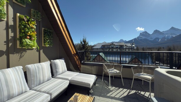 A balcony with a sofa and chairs, a view of mountains, and a building with a green roof in the background.