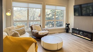 A modern living room with a large window, a sofa, a chair, a round ottoman, and a fish tank.