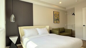 A modern hotel room with a large bed, a side table, a sofa, a painting on the wall, and a coat rack.