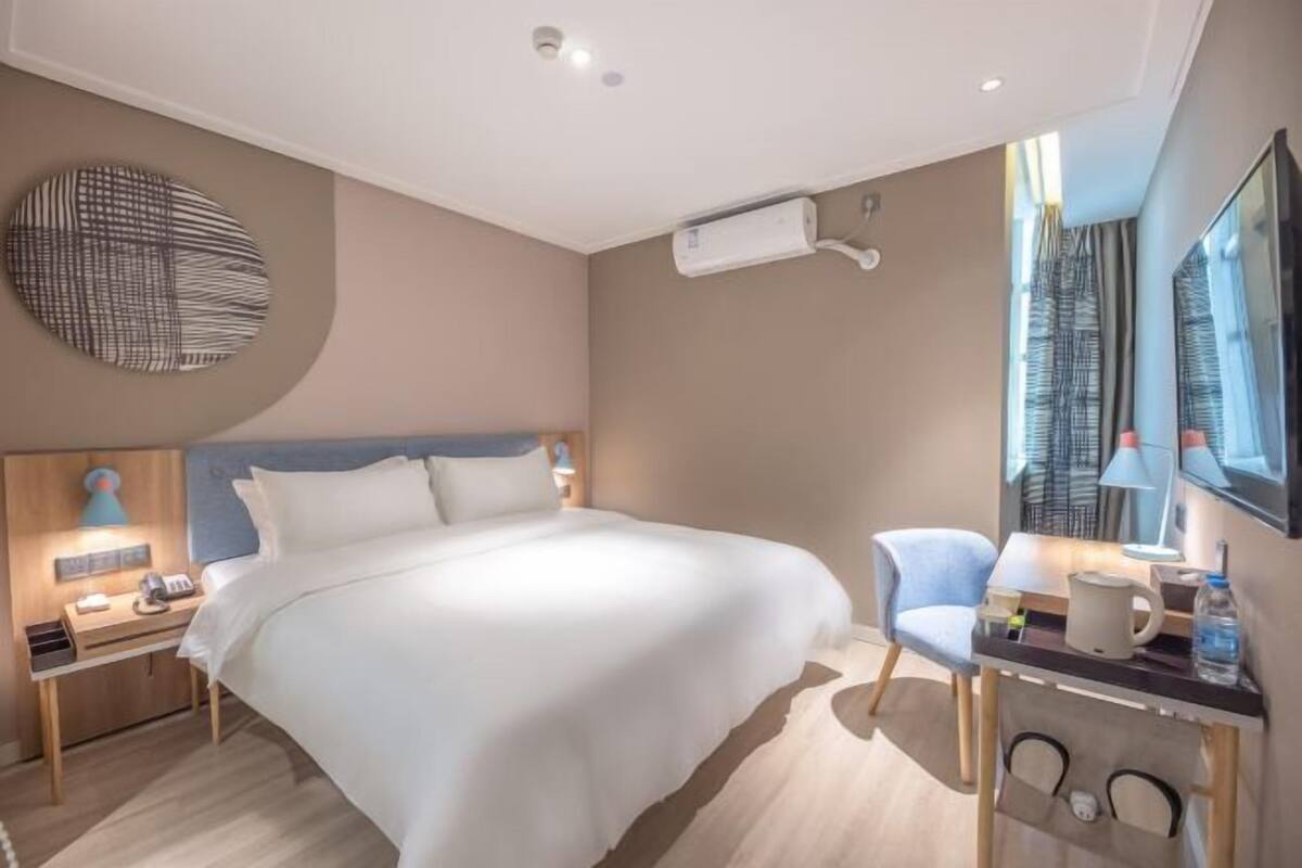 A modern hotel room with a large bed, a desk with a chair, a round wall decoration, and an air conditioning unit.