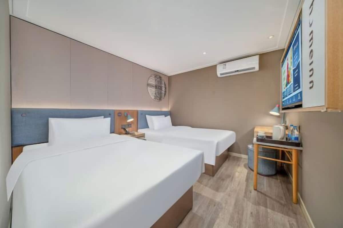 A hotel room with two beds, a small table with a lamp, a trash bin, and an air conditioning unit on the wall.