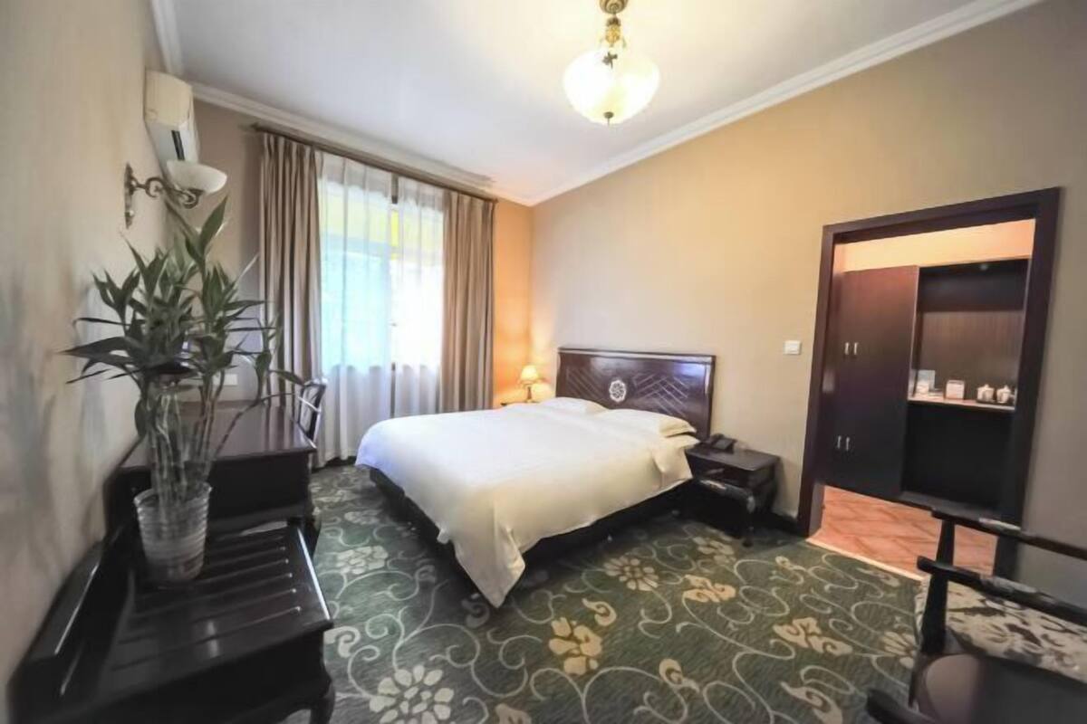 A hotel room with a bed, bedside table, lamp, wardrobe, and a potted plant.
