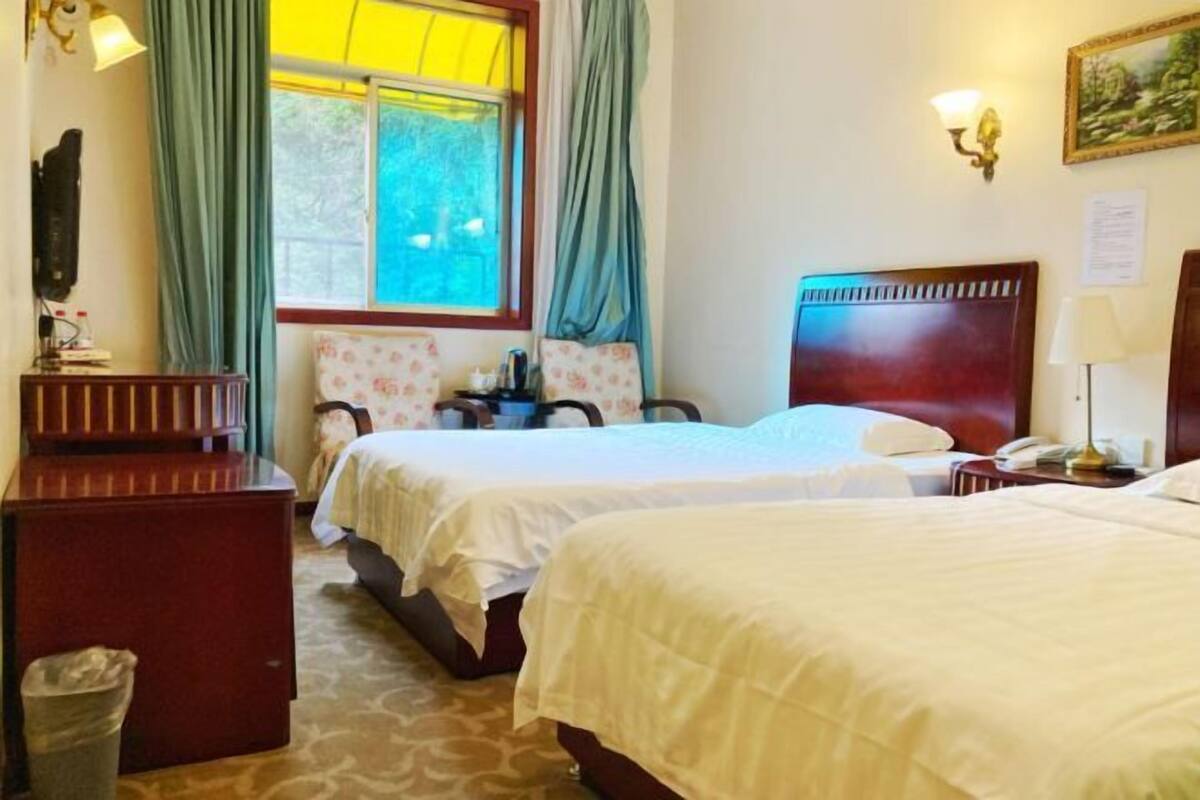 A hotel room with two beds, a desk, a chair, a trash bin, and a window with curtains.