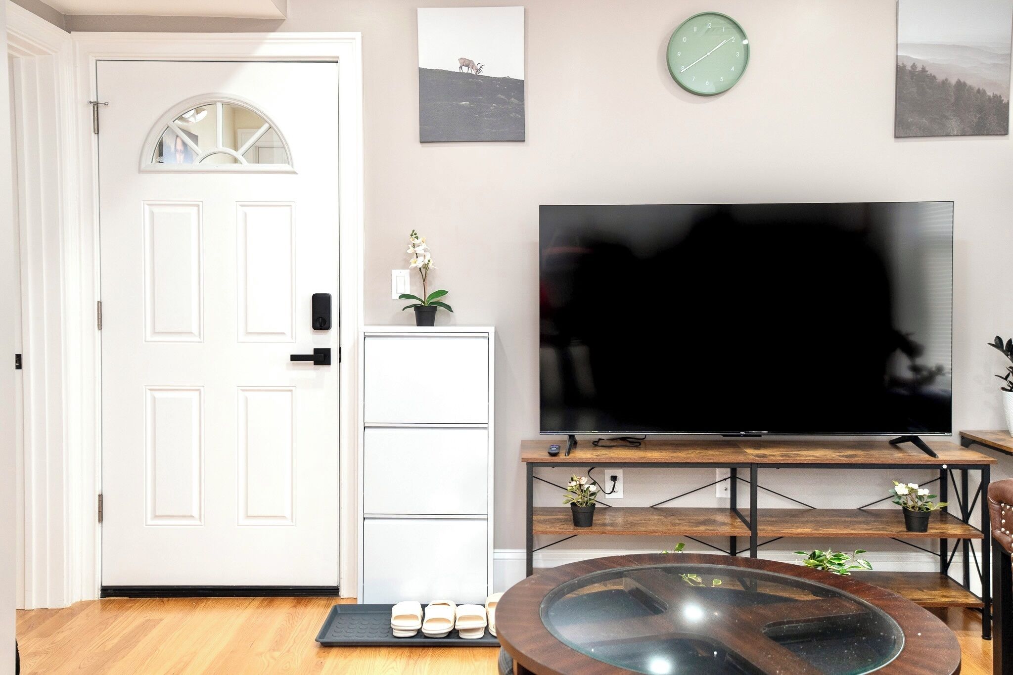 Ein Wohnzimmer mit einer weißen Tür, einem Flachbildfernseher auf einem Holzständer, einem kleinen Topf mit Pflanze und einem Couchtisch mit Glasplatte.