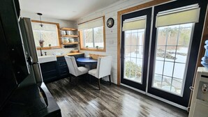 A kitchen with a dining table, chairs, a refrigerator, and a window with blinds. - Escape to nature at Saltwater Hideaway Cottage. Panoramic views!! (Lawrencetown)