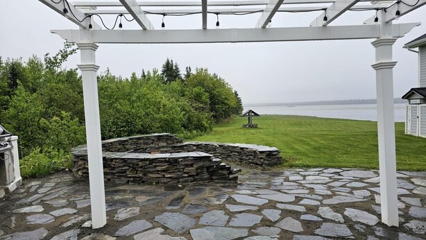 A stone patio with a pergola structure, a stone wall, and a view of a grassy area with a small structure. - Escape to nature at Saltwater Hideaway Cottage. Panoramic views!! (Lawrencetown)