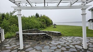 Kivipäällysteinen terassi, jossa on pergola, kivimuuri ja näkymä ruohikkoiselle alueelle, jonka keskellä on pieni rakennelma.