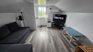 A compact living space with a grey sofa, a small table, a TV, and a window. - Escape to nature at Saltwater Hideaway Cottage. Panoramic views!! (Lawrencetown)