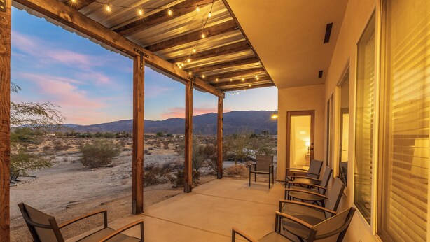 A covered patio with chairs and a table, overlooking a desert landscape with mountains in the distance.