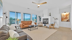 A spacious living room with a brown sofa, a beige armchair, a coffee table, and a large window with blinds.