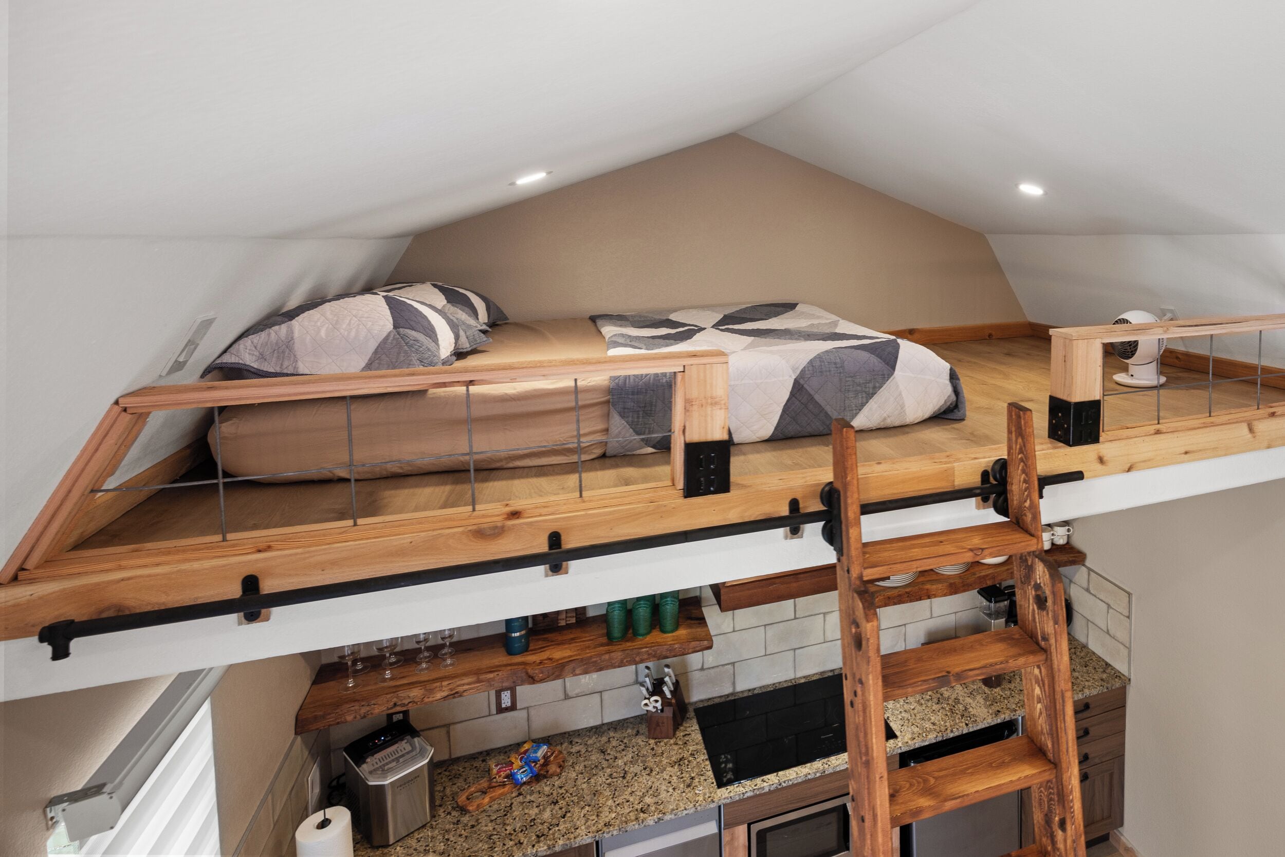 A loft bedroom with a bunk bed, wooden beams, a kitchen area below, and a white ceiling.