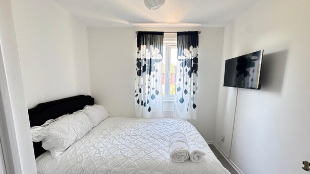 A neatly made bed with a white duvet and rolled towels, a black headboard, a window with black and white curtains, and a flat-screen TV mounted on the wall.