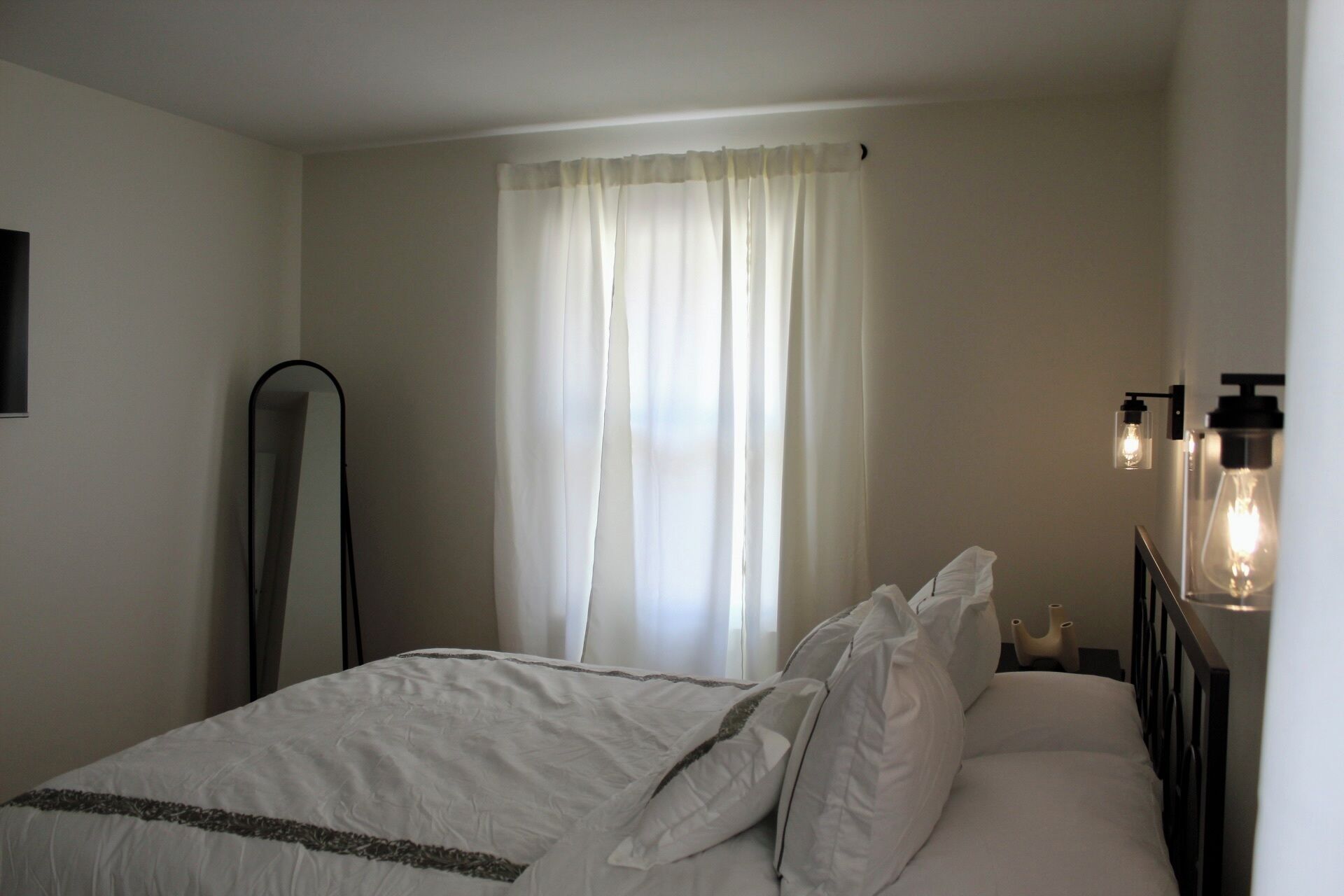 Una camera d'albergo con un letto, tende bianche, uno specchio e luci a parete.