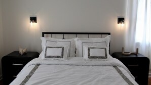 Un lit bien fait, avec une literie blanche et des coussins décoratifs, flanqué de deux tables de chevet surmontées de lampes, dans une pièce simple aux murs unis et dotée d’une fenêtre avec des rideaux.