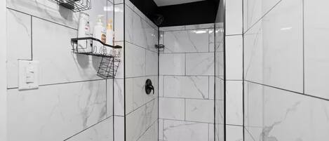 Un baño moderno con azulejos de mármol blanco, zona de ducha y dos toalleros metálicos negros con artículos de aseo.
