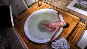 Eine Frau in einer Badewanne in einem Raum mit Fliesenboden, einer hölzernen Sitzbank und einem kleinen Tisch mit einer Flasche und einem Glas.