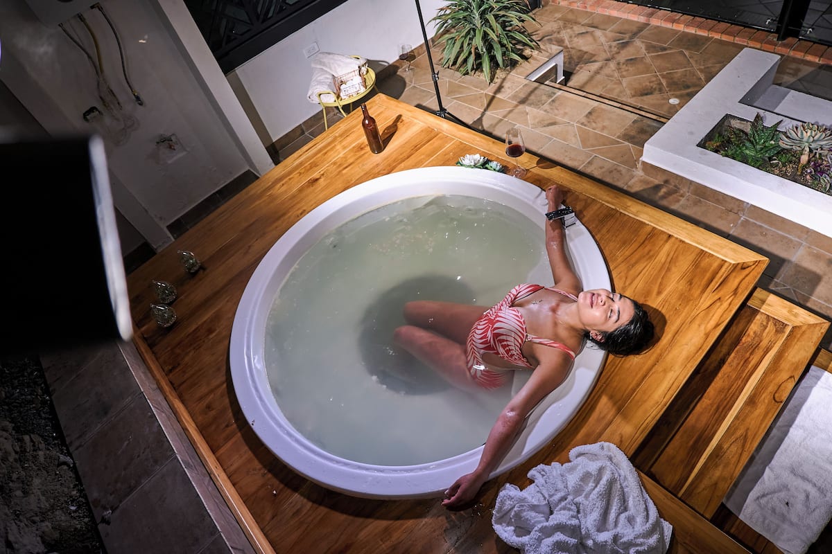 Une femme est dans une baignoire, dans une pièce dont le sol est carrelé, où se trouvent un banc en bois et une petite table sur laquelle reposent une bouteille et un verre.