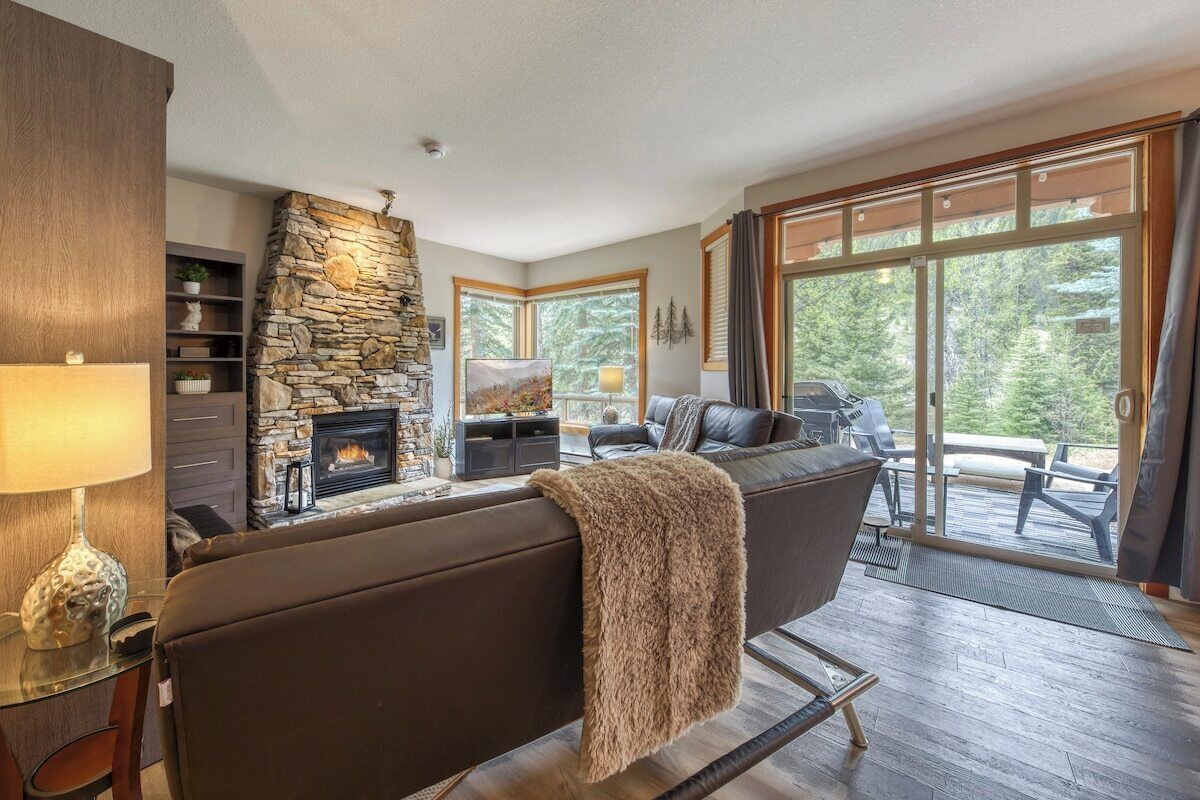 Una sala de estar con chimenea de piedra, un sofá, un televisor y vistas al exterior a través de amplios ventanales.